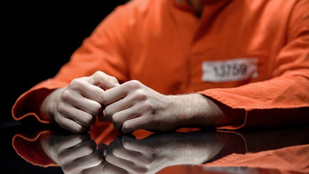 Man in handcuffs and jail jumpsuit sits at a desk